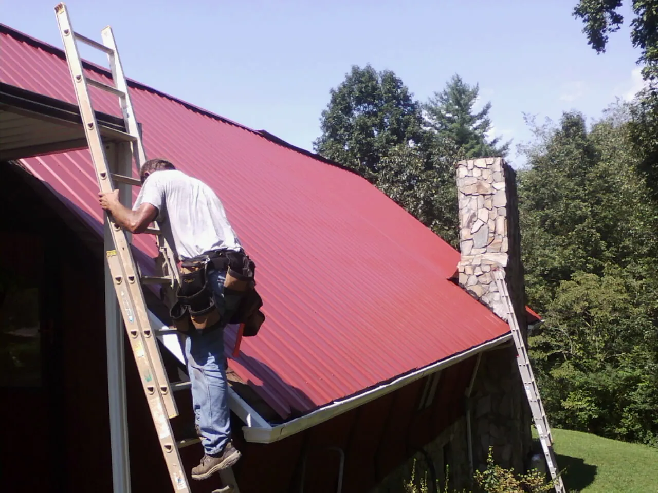 Roofer on a ladder beside a red metal roof and stone chimney — Soffit & Fascia Repair by Roofing Brothers in Tieton